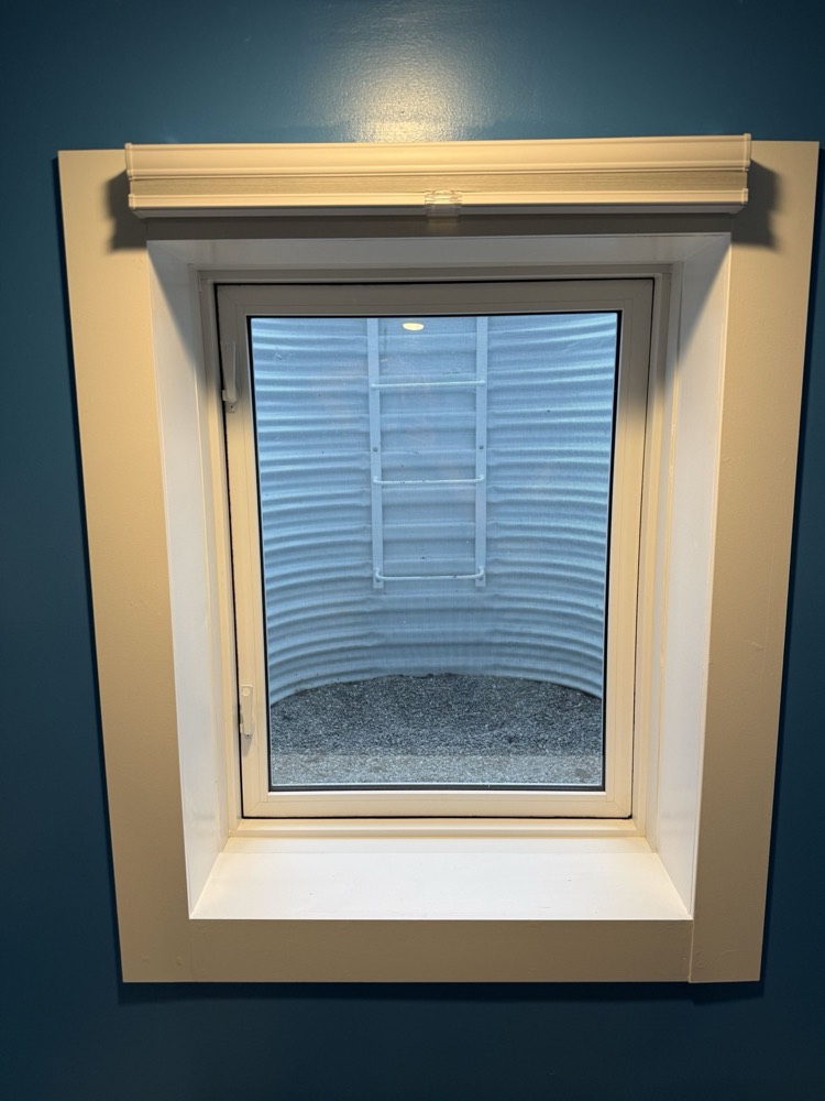 Finished basement egress window with trim and blind in a painted bedroom